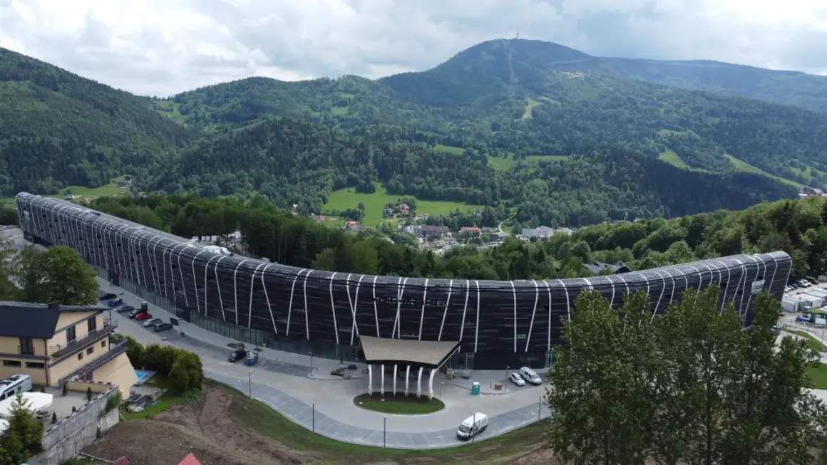 Nowoczesny Mercure Szczyrk Resort w otoczeniu zielonych gór. Budynek o falistym kształcie z czarną fasadą i białymi elementami.