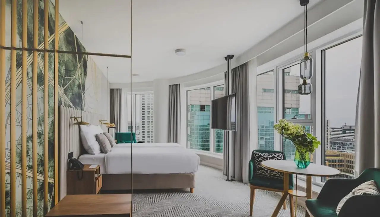 Elegancki pokój hotelowy z widokiem na miasto. Dwa łóżka, stolik z krzesłami i telewizor. Idealne miejsce na pobyt w Royal Tulip Warszawa.