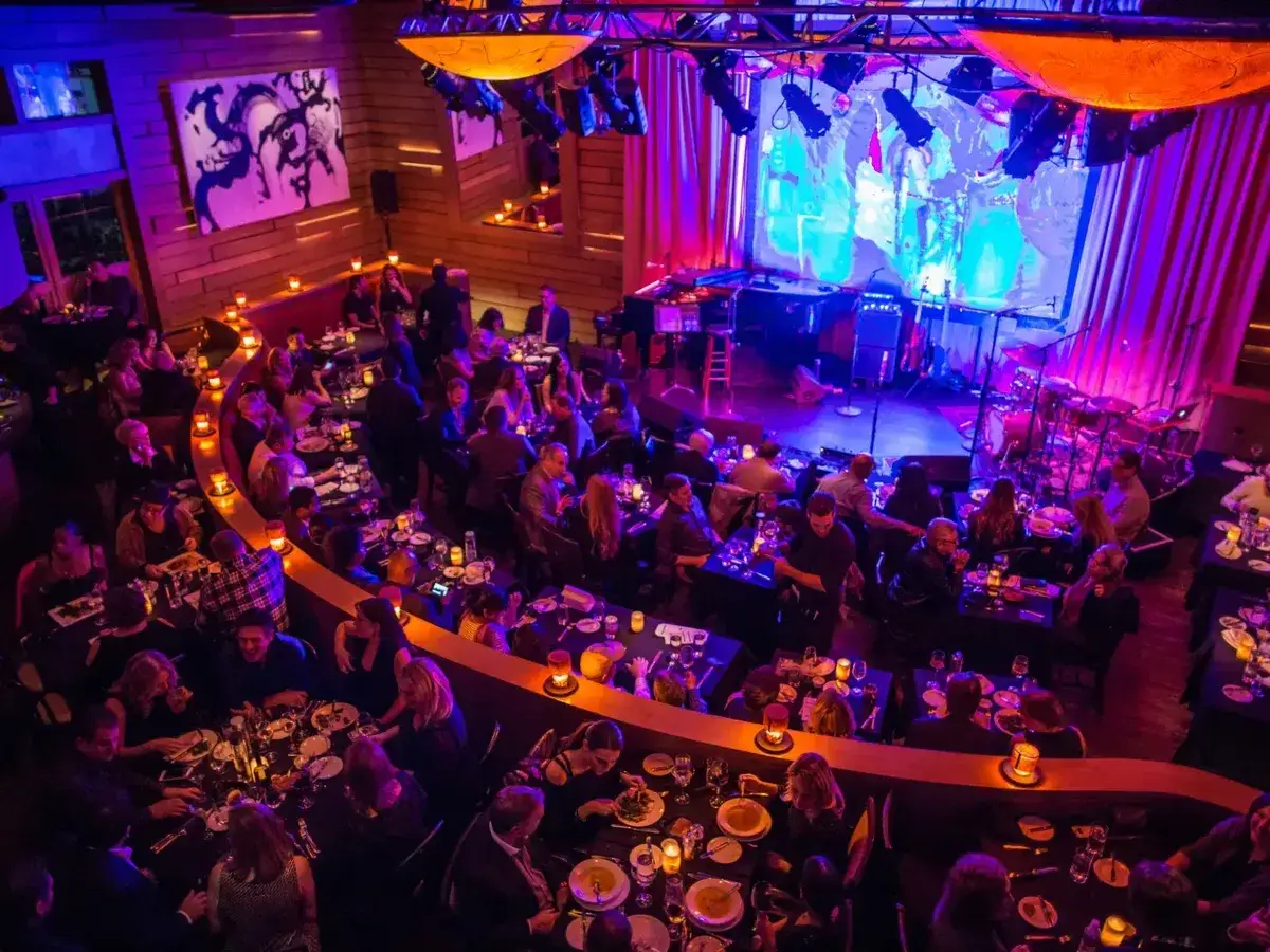 Elegancka restauracja z muzyką na żywo w Warszawie. Goście siedzą przy stolikach, delektując się posiłkami i atmosferą. Na scenie przygotowany sprzęt muzyczny.